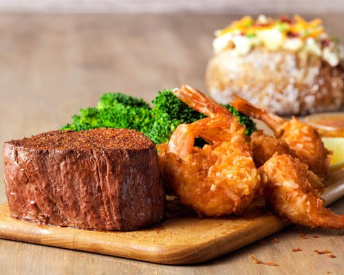 A wooden platter featuring a seasoned beef filet, crispy shrimp, and steamed broccoli, with a baked potato in the background.