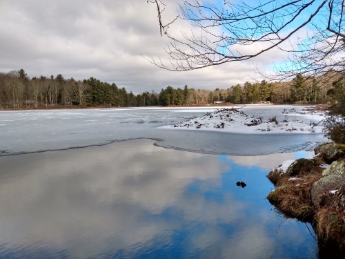 A serene winter landscape featuring a frozen lake, snow-covered banks, and trees reflecting in the water.