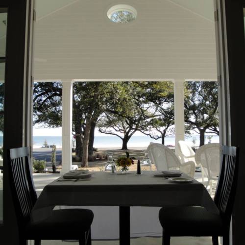 A view from inside a dining area, featuring a table set for two and a scenic outdoor view of trees and the ocean.