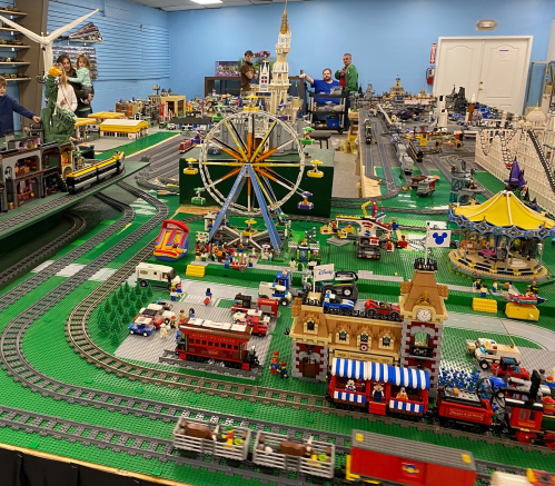 A vibrant model train display featuring a carnival, buildings, and a Ferris wheel, with people observing in the background.