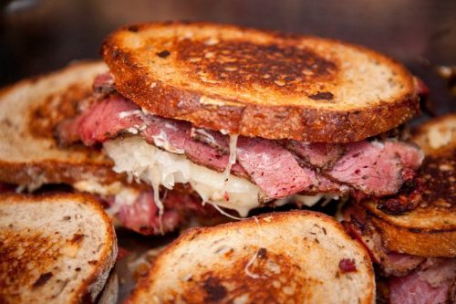 A close-up of a toasted sandwich filled with layers of pastrami and melted cheese, with sauerkraut peeking out.