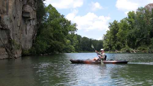 A person kayaking on a calm river surrounded by lush greenery and rocky cliffs under a blue sky.