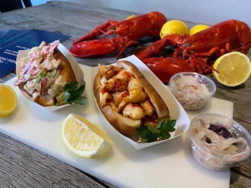 Two lobster rolls with sides of coleslaw, fresh lemons, and whole lobsters in the background on a wooden table.