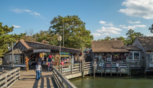A wooden boardwalk leads to shops by a calm waterway, with people browsing and enjoying a sunny day.