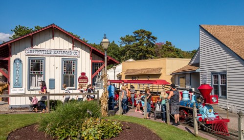 A historic railroad station with a steam engine, surrounded by people and quaint buildings in a charming village setting.
