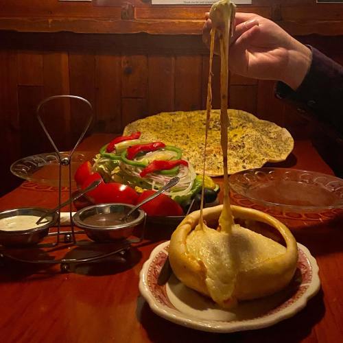 A hand pulls a cheesy dish from a bread bowl, with a salad and flatbread in the background on a wooden table.