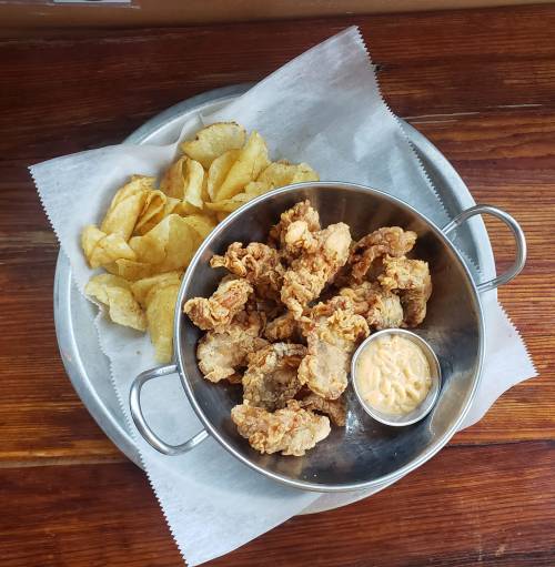 A metal bowl filled with crispy fried chicken pieces and a small cup of dipping sauce, accompanied by potato chips.