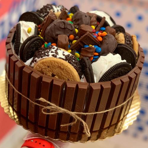 A decadent cake topped with cookies, chocolate candies, and various sweets, surrounded by Kit Kat bars.