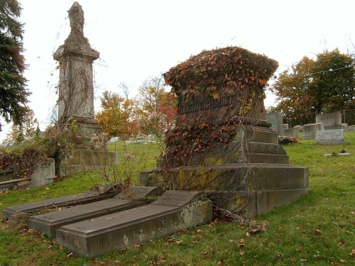 A weathered gravestone covered in vines, surrounded by a cemetery with autumn foliage in the background.