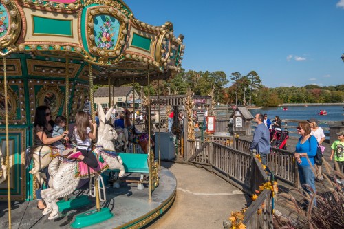 A colorful carousel with unicorns, people enjoying the park, and a lake in the background on a sunny day.