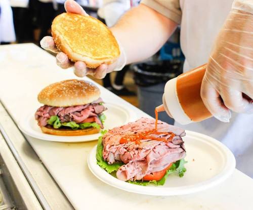 A hand adds ketchup to a roast beef sandwich on a plate, with another sandwich and a bun nearby.