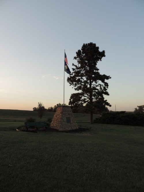 A stone monument with a flag stands in a grassy area, beside a tall tree, under a clear sky at sunset.