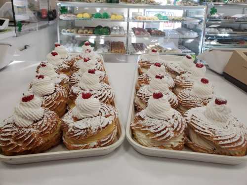 Two trays of pastries topped with whipped cream and cherries, displayed in a bakery with various sweets in the background.