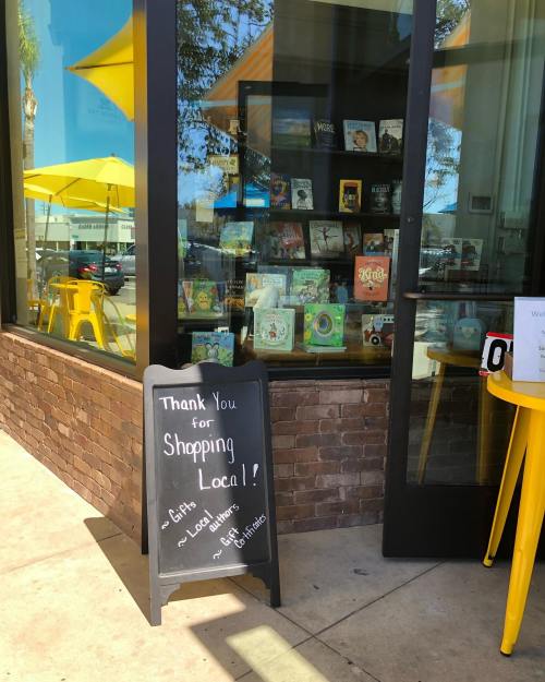 A storefront with a sign thanking customers for shopping local, featuring colorful books displayed in the window.