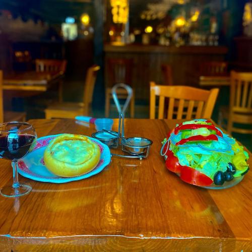 A wooden table with a glass of red wine, a bread bowl filled with soup, and a colorful salad on a plate.