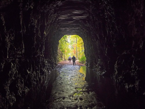 A dark tunnel with a wet floor, leading to a bright, colorful forest in the distance, with two figures walking towards the light.