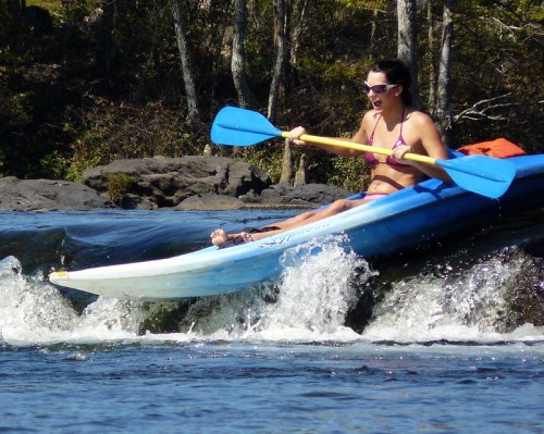 A woman in a bikini kayaks over a small wave, surrounded by trees and rocky shoreline.