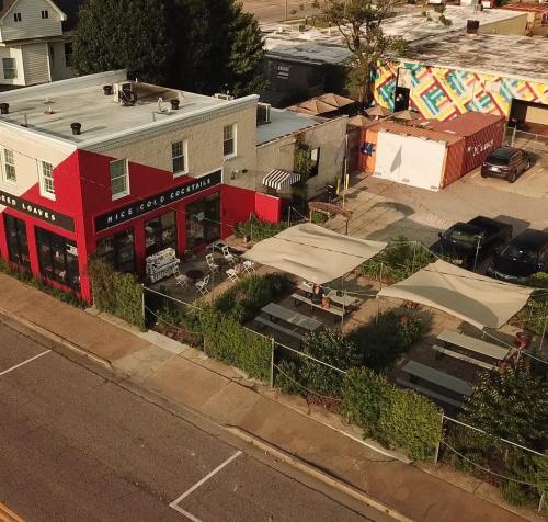 Aerial view of a red building with outdoor seating, surrounded by greenery and parked cars on a sunny day.