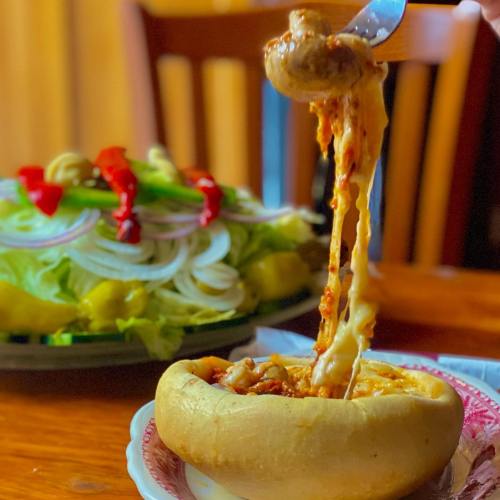 A cheesy pasta dish in a bread bowl, with a fresh salad in the background, featuring lettuce, onions, and peppers.