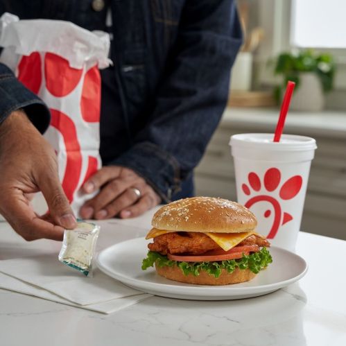 A hand reaches for a sauce packet next to a chicken sandwich on a plate, with a drink and a takeout bag in the background.