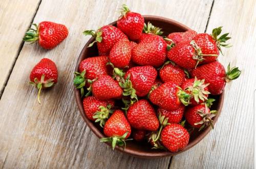 A wooden bowl filled with fresh, ripe strawberries on a rustic wooden surface.