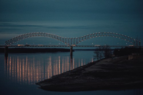 A lit bridge arches over a river at dusk, reflecting lights on the water's surface.