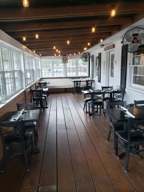 A cozy restaurant interior with wooden floors, black tables, and string lights, featuring large windows and ceiling fans.