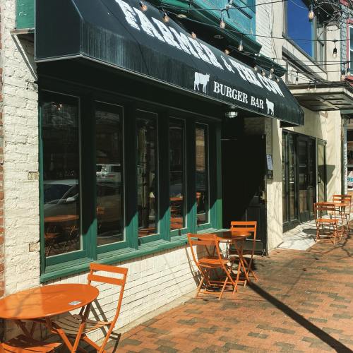 Exterior of a burger bar with green windows and orange tables on a brick sidewalk, under a black awning.
