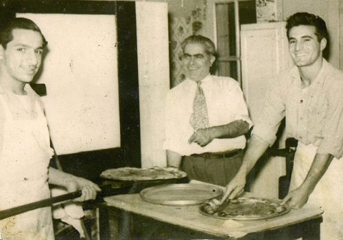 Three men in a kitchen, one holding a pizza on a peel, another with a pizza on a tray, and a third smiling in the background.