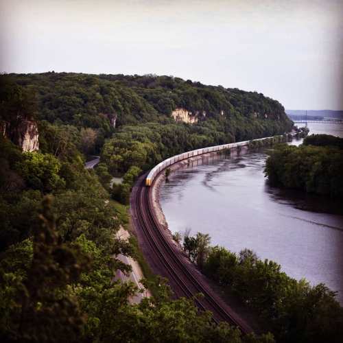 A train curves along a riverbank, surrounded by lush green hills and trees under a soft, overcast sky.