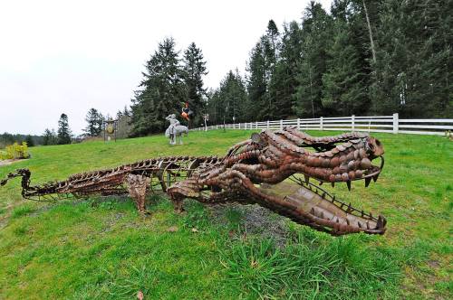 A large, rusted metal sculpture of a two-headed dragon lies on green grass, surrounded by trees and a white fence.