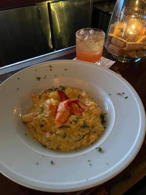A bowl of creamy lobster risotto garnished with herbs, alongside a cocktail in a dimly lit setting.