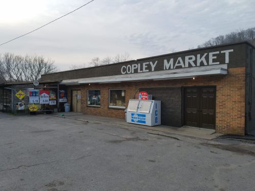 Exterior of Copley Market, a small store with a sign, parking area, and vending machines outside. Trees in the background.