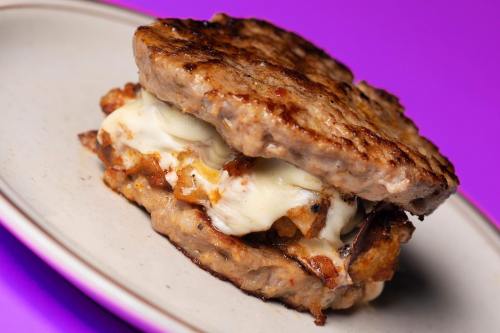 A hearty sandwich made with two sausage patties, filled with melted cheese and crispy bits, on a vibrant purple background.
