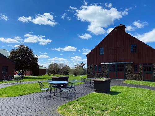 A sunny outdoor scene featuring a red barn, stone building, and metal tables on a grassy area under a blue sky.