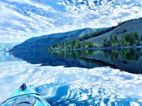 A serene lake scene with a kayak in the foreground, reflecting clouds and mountains under a blue sky.