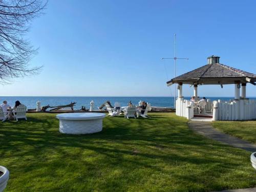 A serene lakeside scene with a gazebo, white chairs, and a clear blue sky. People relax by the water's edge.