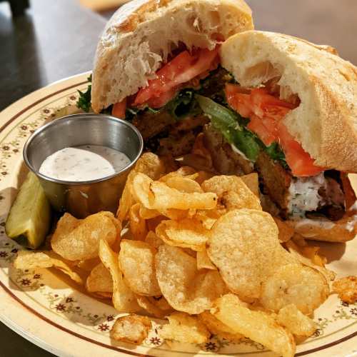 A plate with a sandwich filled with lettuce and tomato, served with chips and a small cup of dipping sauce.