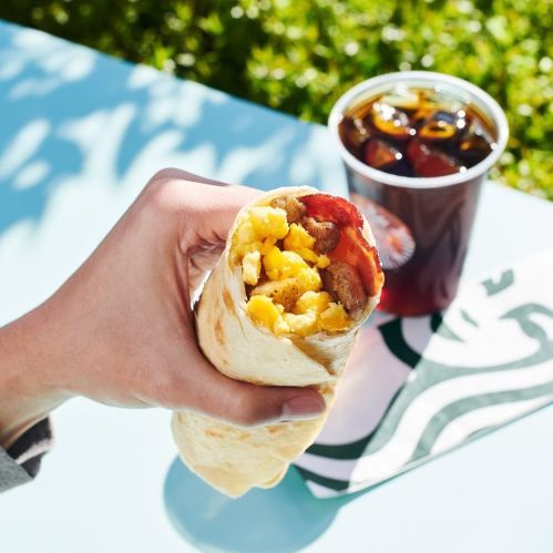 A hand holding a breakfast burrito filled with eggs and bacon, next to a cup of iced coffee on a table.