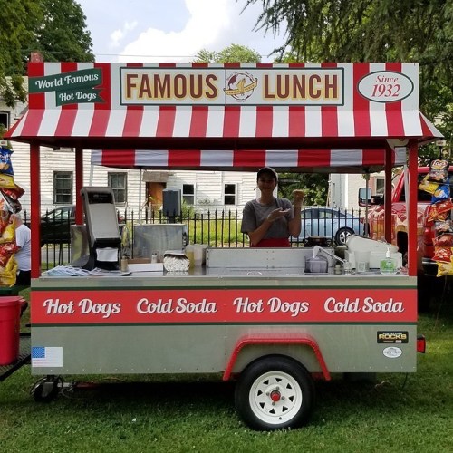 A food cart named "Famous Lunch" selling hot dogs and cold soda, with a vendor preparing food.