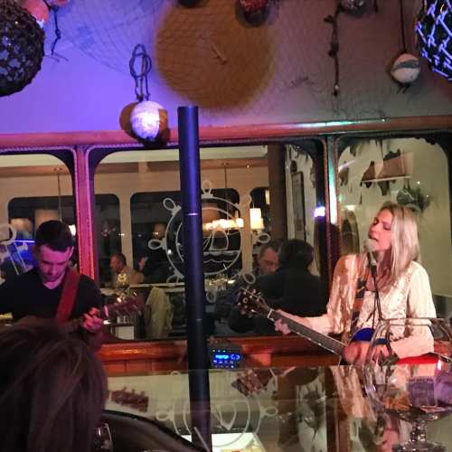 A musician performs on stage with a guitar, while another musician plays a mandolin in a cozy venue.
