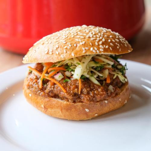A sesame seed bun filled with savory meat and topped with a colorful slaw of carrots and cabbage.