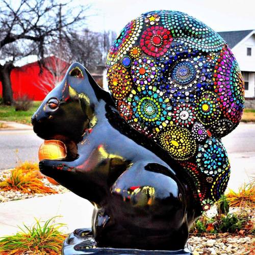 A colorful, mosaic-patterned squirrel statue holding an acorn, set against a blurred background of grass and buildings.