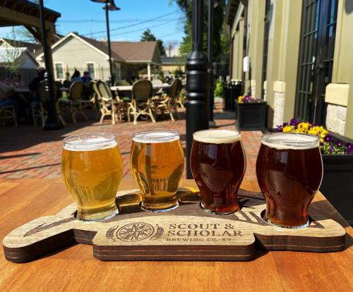 A wooden flight board holds four craft beer glasses in a sunny outdoor setting, with tables and flowers in the background.