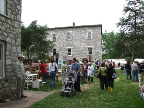 A crowd gathers outside a stone building, browsing items for sale at a local market or fair on a cloudy day.