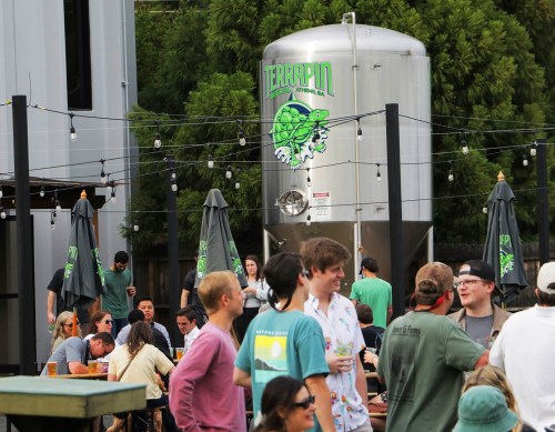 A lively outdoor gathering at a brewery, with people socializing around tables and a large Terrapin Brewing tank in the background.