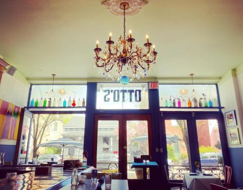 A bright restaurant interior featuring a chandelier, colorful bottles on a shelf, and large windows with outdoor seating.