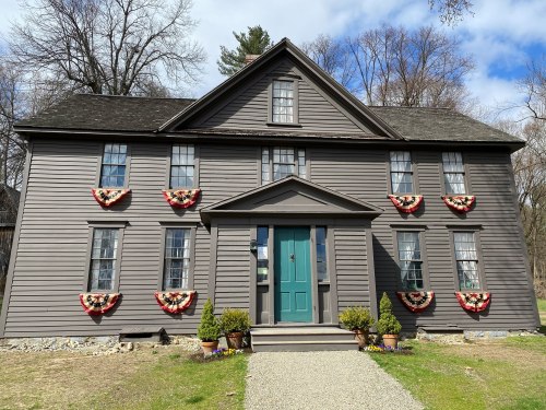 A gray house with a green door, decorated with floral wreaths, surrounded by trees and a gravel path.