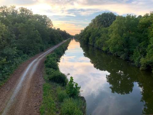 A serene canal lined with trees, reflecting the sky at sunset, with a dirt path alongside.