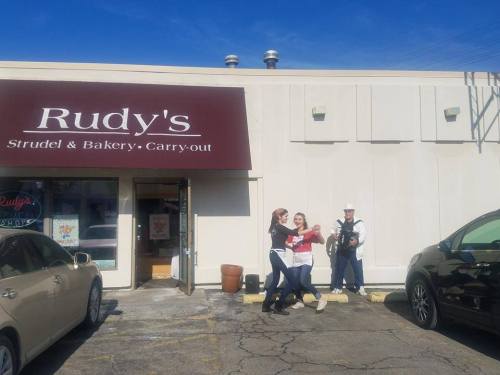 A bakery storefront with a sign reading "Rudy's Strudel & Bakery." Two people pose outside, while another plays an accordion.